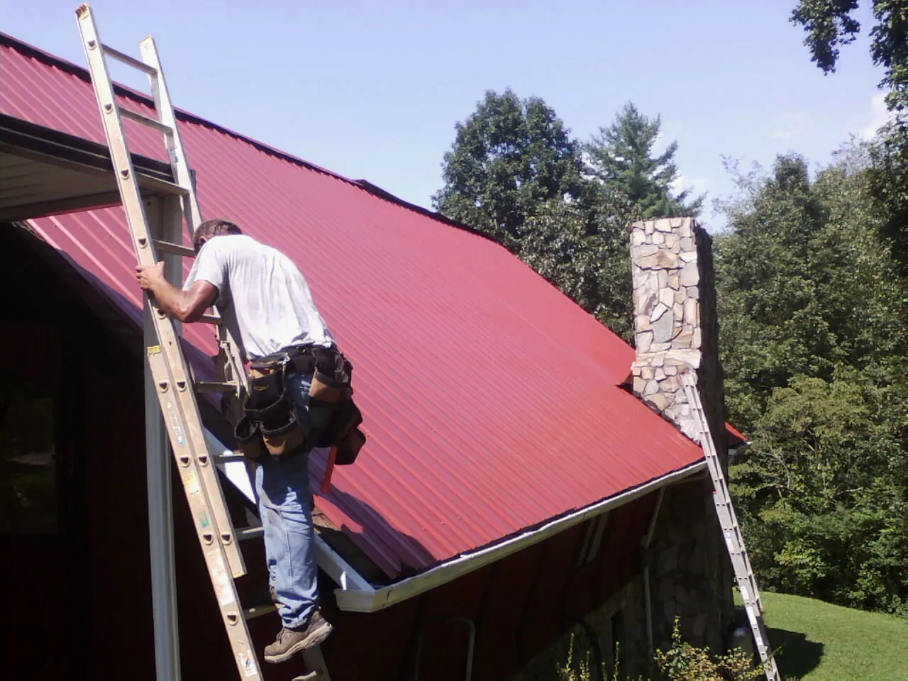 Roofer on a ladder beside a red metal roof and stone chimney — Soffit & Fascia Repair by Roofing Brothers in Randalia